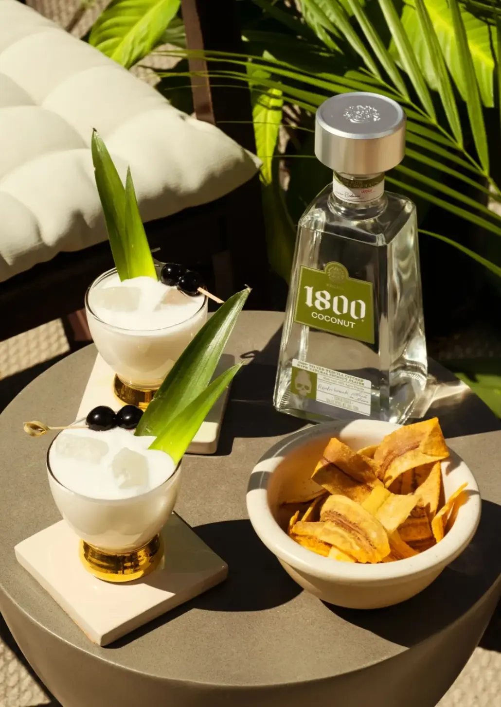 bottle of 1800 coconut, 2 rocks glasses of coconut margarita and a bowl of plantain chips